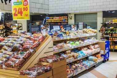 Market pastanesi bölümündeki ahşap raflarda, parlak tanıtım tabelalarıyla çeşitli paketlenmiş ekmek çeşitleri.