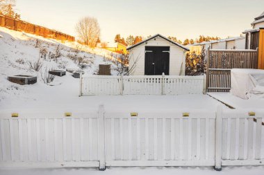 Beyaz ahşap çitli, karla kaplı arka bahçesi, küçük bir bahçe kulübesi ve kış günü yükseltilmiş bahçe yatakları. İsveç.