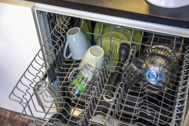 Temiz cam eşyalar, seramik bardaklar ve metal raflarda düzgünce dizilmiş yeşil tabaklarla dolu açık bir bulaşık makinesi..