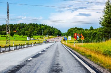 Yeşil tarlalar, trafik işaretleri ve orman ile çevrili düz asfalt yol parçalı bulutlu gökyüzü altında. 