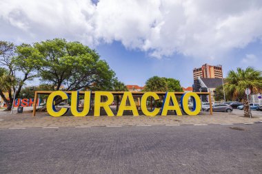 Büyük sarı Curacao tabelaları ağaçlar, binalar ve arka planda park edilmiş arabalarla açık havada sergileniyordu.. 