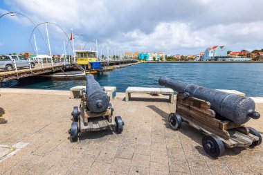 Willemstad 'daki Kraliçe Emma Köprüsü' ne bakan tarihi toplar. Arka planda renkli deniz manzaralı binalar var.. 