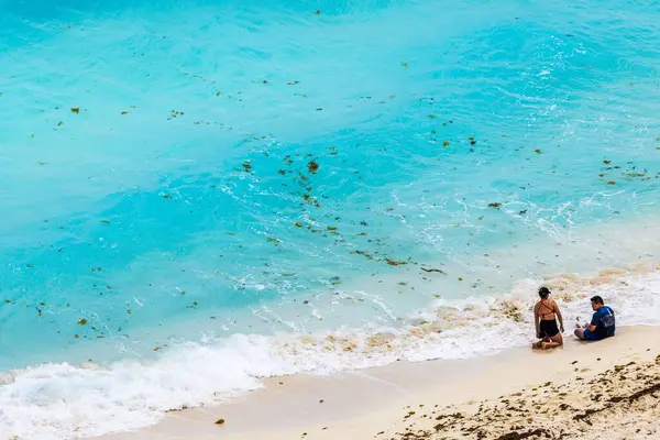 Turkuaz Karayip Denizi dalgaları ve ön planda deniz yosunlarıyla kumlu sahilde oturan çift. Cancun..