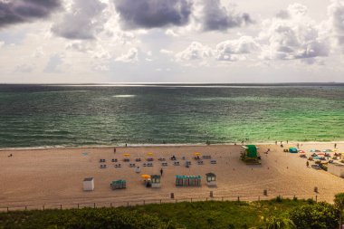 Zümrüt yeşili Atlantik Okyanusu manzarası. Dalgalar kumlu sahile ulaşıyor. Güneş panjurları ve bulutlu gökyüzünün altında renkli şemsiyeler var. Miami Plajı. ABD. 