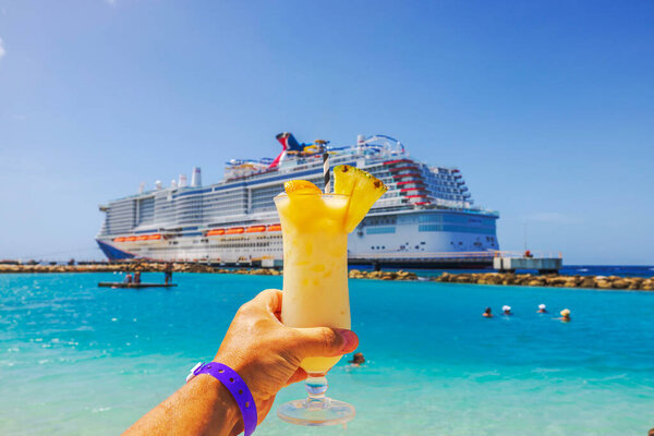 Hand holding alcohol tropical cocktail with pineapple and orange against turquoise water Caribbean Sea and cruise ship. Curacao.
