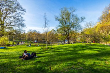 İnsanlar Central Park 'ta yeşil çimlerde ağaçlarla, yürüyüş yollarıyla ve arka planda göletle güneşli bir bahar gününde dinleniyorlar. - New York. ABD