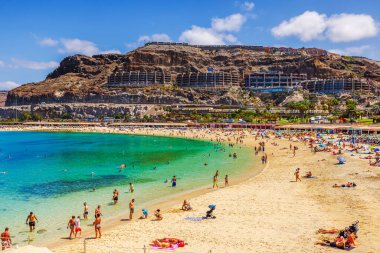 Turkuvaz sularının ve güneşlenmenin tadını çıkaran turistlerle dolu kalabalık kumlu plajlar, otelleri olan kayalık kayalıklar tarafından destekleniyor. Gran Kanaryası, Las Palmas, İspanya. 