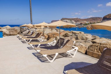 Bej şemsiyeler altında boş güneş panzeleri. Atlantik Okyanusu 'na bakan kayalık sahil şeridi ve arka planda kayalıklar. Gran Kanaryası, Las Palmas, İspanya.