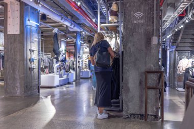 Back view of woman browsing sports apparel in Adidas store with modern industrial interior and stylish clothing displays. New York. USA.  clipart