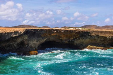 Dalgalar Arikok Ulusal Parkı 'ndaki doğal mağara yakınlarındaki kayalık sahil şeridine çarpıyor. Mavi gökyüzünün altında bulutlar var. Aruba.