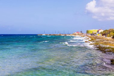 Turkuaz dalgalı Karayip Denizi kıyı manzarası, kayalık kıyı şeridi, açık gökyüzü altında küçük iskele ve renkli binalar. Curacao.