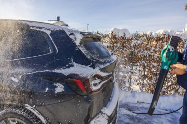Adam, güneşli bir kış günü banliyö evinin önüne park etmiş siyah SUV 'den kar temizlemek için elektrikli hava üfleyici kullanıyor. İsveç.