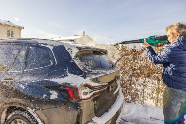 Adam, güneşli bir kış günü banliyö evinin önüne park etmiş siyah SUV 'den kar temizlemek için elektrikli yaprak üfleyicisi kullanıyor. İsveç.