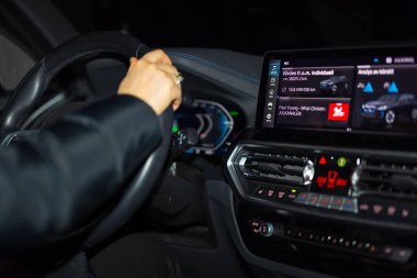 Gece BMW iX3 elektrikli aracın direksiyonunu tutan sürücünün yakın görüntüsü. Işıklı gösterge paneli ve dokunmatik ekran. İsveç.