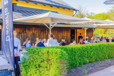 Güneşli bir günde, kırsal ahşap binanın yakınındaki büyük güneş gözlüğü altında yemek yiyen insanların olduğu açık hava restoran terası. İsveç.