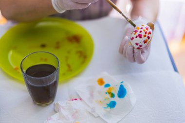 Paskalya yumurtasını kırmızı ve turuncu çiçekli elle boyamak için beyaz masadaki fırçayı sanat malzemeleriyle birlikte kullanın. İsveç.