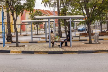 Bulutlu bir günde otobüs durağında metal barınaklar, banklar, ağaçlar ve renkli binalarla oturan iki kişi. Willemstad. Curacao.