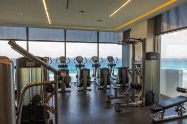 Antrenman ekipmanları ve Karayip Denizi kıyı şeridine bakan panoramik pencereli modern fitness salonu. Meksika mı? Cancun..