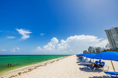 Miami sahilinde kumsalda mavi şemsiyeler altında dinlenen ve Atlantik Okyanusunda yüzen turistler var. Miami Plajı. ABD.
