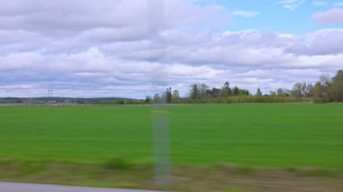 Yeşil tarlaları, orman çizgileri ve hareket halindeki araçtan görülen bulutlu gökyüzü olan bahar tarlaları. İsveç.