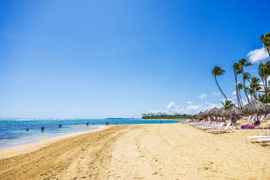Atlantik Okyanusunda yüzen ve kumlu sahildeki palmiye kaplı şemsiyelerin altında şezlonglarda dinlenen turistler. Punta Cana. Dominik Cumhuriyeti.