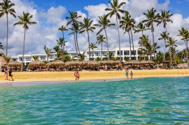İnsanlar kumlu sahilde palmiye ağaçları, sazdan şemsiyeler ve Atlantik Okyanusu kıyısındaki otelle yürüyüp rahatlıyorlar. Punta Cana. Dominik Cumhuriyeti.