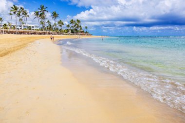 Turistler, zümrüt Atlantik Okyanusu ve mavi bulutlu gökyüzünün altında palmiye ağaçları olan kumlu bir plaj. Punta Cana. Dominik Cumhuriyeti. 