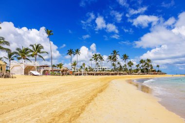 Atlantik Okyanusu 'nun kumlu sahili. Palmiye ağaçları, beyaz kabinler ve mavi gökyüzünün altında dalgalar. Punta Cana. Dominik Cumhuriyeti.