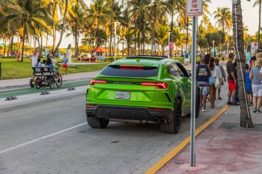 Yeşil Lamborghini Urus, gün batımında turistler ve palmiye ağaçlarıyla çevrili Miami 'deki Ocean Drive' a park etmiş. Miami Plajı. ABD. 