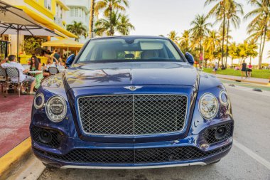 Miami Plajı 'ndaki Ocean Drive' a park etmiş mavi Bentley Bentayga SUV 'nin ön görüntüsü. Palmiye ağaçları ve arka plandaki açık kafede insanlar var. Miami Plajı. ABD. 