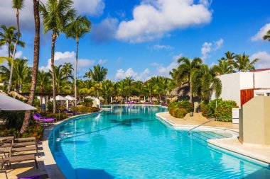 Parlak mavi gökyüzünün altındaki lüks bir tatil köyünde, palmiye ağaçları ve şezlonglarla çevrili tropik yüzme havuzu. Punta Cana. Dominik Cumhuriyeti. 