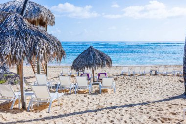 Atlantik Okyanusu ve bulutlu gökyüzü ile parlak kumlu sahilde saman şemsiyelerin altında beyaz şezlonglar. Dominik Cumhuriyeti. Punta Cana. 