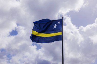 Curacao ulusal bayrağı bulutlu gökyüzüne karşı uzun bayrak direğinde dalgalanıyor ve güneş ışığı bulutları delip geçiyor. Willemstad. Curacao.