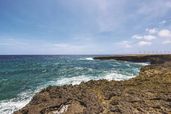 Kayalık kıyı şeridi, Karayip Denizi dalgaları güneşli gökyüzünün altına vuruyor ve arka planda rüzgar türbinleri var. Aruba.
