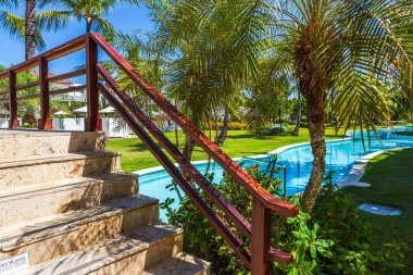 Güneşli tropik tatil köyü bahçesindeki palmiye ağaçları arasında kavisli havuzun üzerinde ahşap korkuluklu taş merdivenler. Dominik Cumhuriyeti. Punta Cana. 