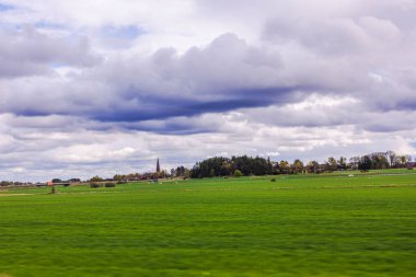 İsveç kırsalında, dramatik bulutlu gökyüzü altında uzak kilise kulesiyle yeşil bahar tarlaları.