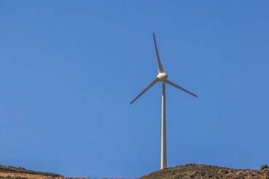Açık mavi gökyüzünün altında, dağın tepesinde duran rüzgar türbini güneşli havada yenilenebilir enerji üretiyor. Kos. Yunanistan.