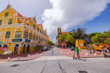 İnsanlar Penha binası ve şehir saati yakınlarındaki rıhtım boyunca scooterla geziyorlar. Willemstad. Curacao. 08.05.2025.