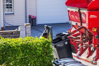 İşçi, güneşli banliyö villa bölgesinde siyah çöp kutusunu kırmızı çöp kamyonuna boşaltıyor. 