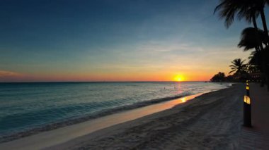 Kumlu Kartal Sahili ve palmiye ağaçlarıyla otel bölgesinden gün batımında Karayip Denizi manzarası. Aruba.