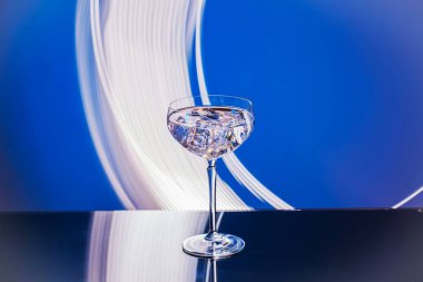 Soyut mavi arkaplana karşı yansıtıcı yüzeyde şeffaf içecek ve buz küpleriyle dolu zarif cam görüntüsünü kapat.