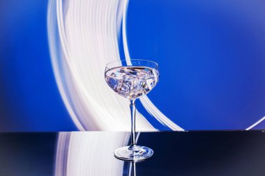 Soyut mavi arkaplana karşı yansıtıcı yüzeyde şeffaf içecek ve buz küpleriyle dolu zarif cam görüntüsünü kapat.