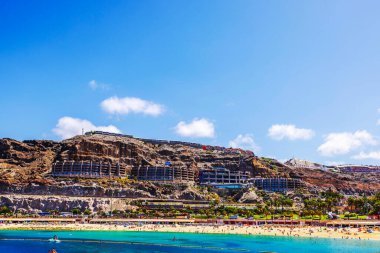 Atlantik Okyanusu 'nda kayalık uçurumlar ve otellerle yüzen turistlerle dolu kumsal. Arka planda bulutlu mavi gökyüzü. İspanya. Büyük Kanarya. 
