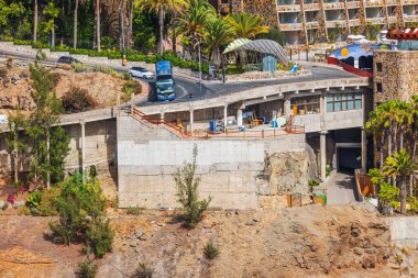 Kayalık yamaçta tamamlanmamış beton bir bina. Arka planda palmiye ağaçları ve turist otobüsü var. İspanya. Büyük Kanarya. 