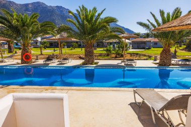 Arka planda palmiye ağaçları ve dağlarla çevrili temiz suyla otel avlusundan yüzme havuzuna manzara. Yunanistan. Kos. 