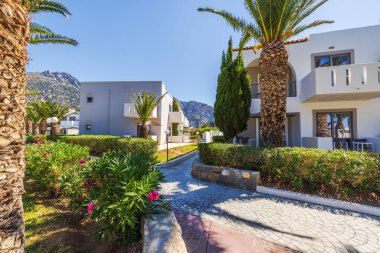 Palmiye ağaçları, yeşil çalılar ve yaz güneşinde dağ manzaralı balkonları olan tatil evleri. Kos. Yunanistan.