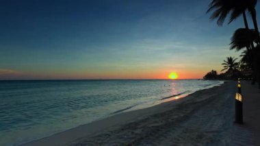 Karayip Denizi üzerinde gün batımı manzarası kumlu plaj, palmiye ağaçları ve renkli gökyüzüne karşı sakin su. Aruba.