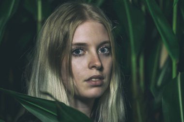 Uzun saçlı esmer kadının korkunç portresi yeşil bitkiler arasında poz veriyor.