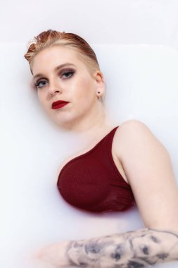 Mavi gözlü, kırmızı dudaklı ve dövmeli çekici genç bir kadın kaplıca banyosunda sütle kameraya bakıyor. Dikey. 