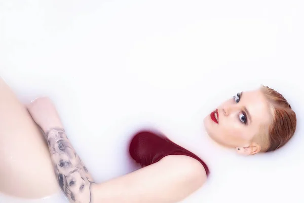 Attractive young woman with blue eyes, red lips and tattoo is lying in spa bath with milk and looking at camera. Horizontally.